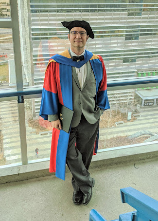 Tyson, smiling, in UCalgary PhD Robes, in stairs overlooking the University of Calgary.